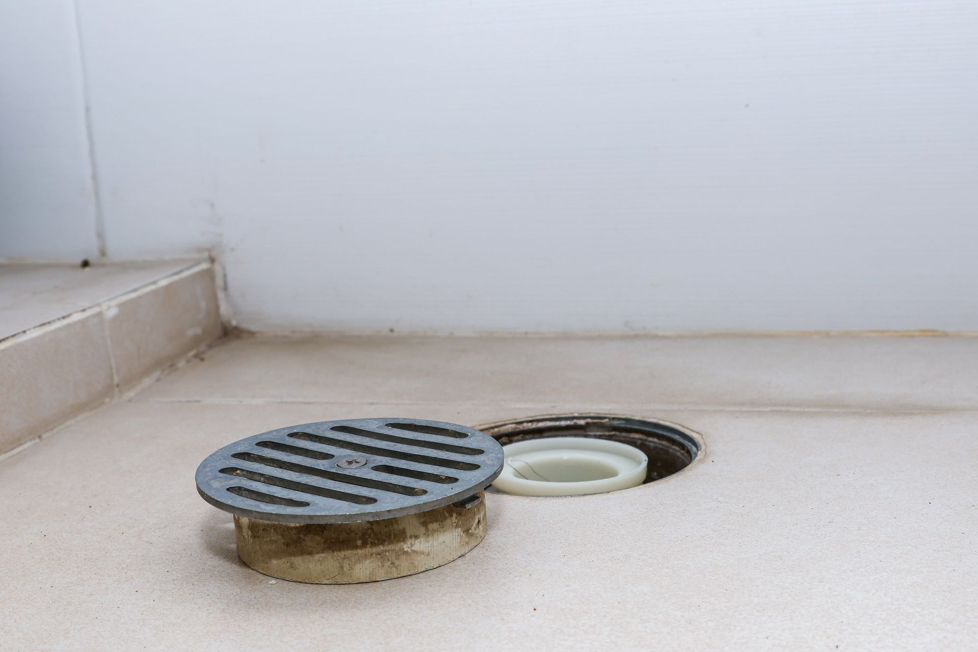 Close-up of a shower drain with a removable, circular metal grate on a tiled floor. Another drain opening is visible.