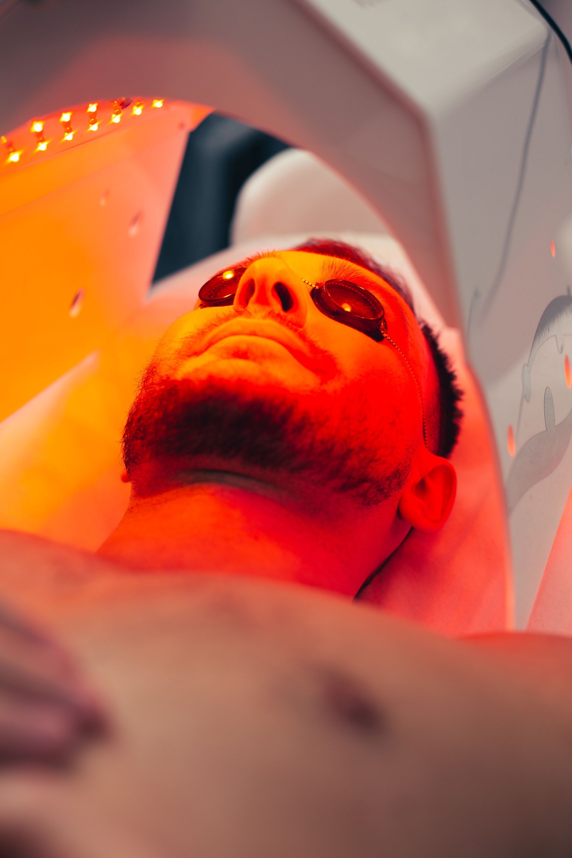 Man undergoing red light therapy, wearing eye protection. Orange glow illuminates his face.