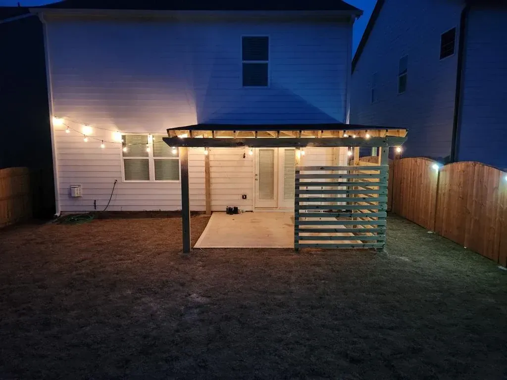 Backyard patio at dusk, with string lights illuminating the space. A wooden pergola and fence are visible.