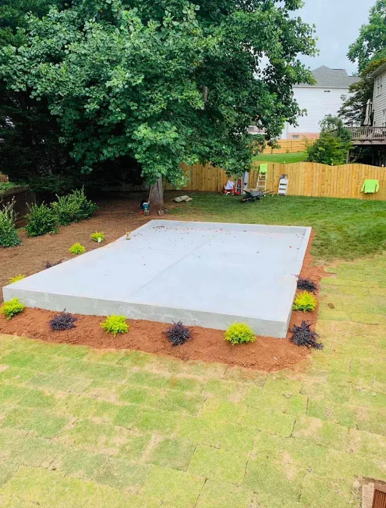 Concrete patio in a backyard, surrounded by bushes and mulch; a tree shades the area.