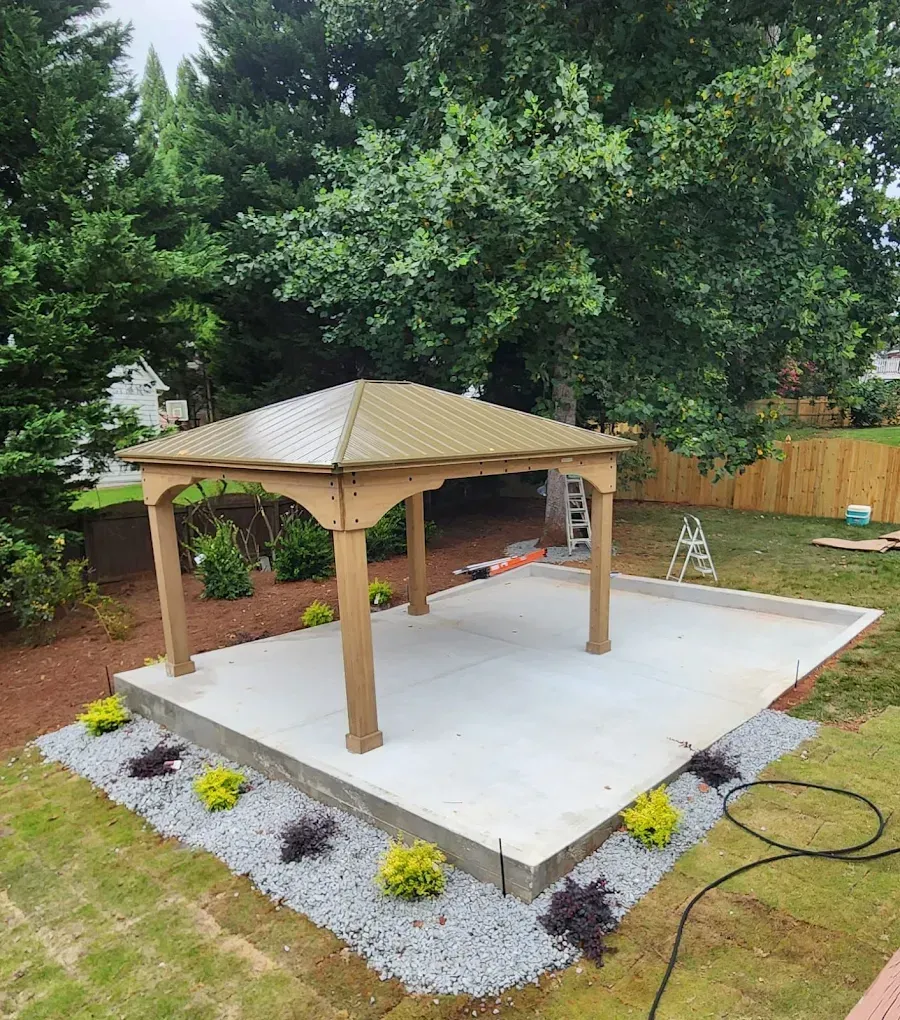 Gazebo on concrete patio, surrounded by gravel, bushes, and grass; backyard setting.