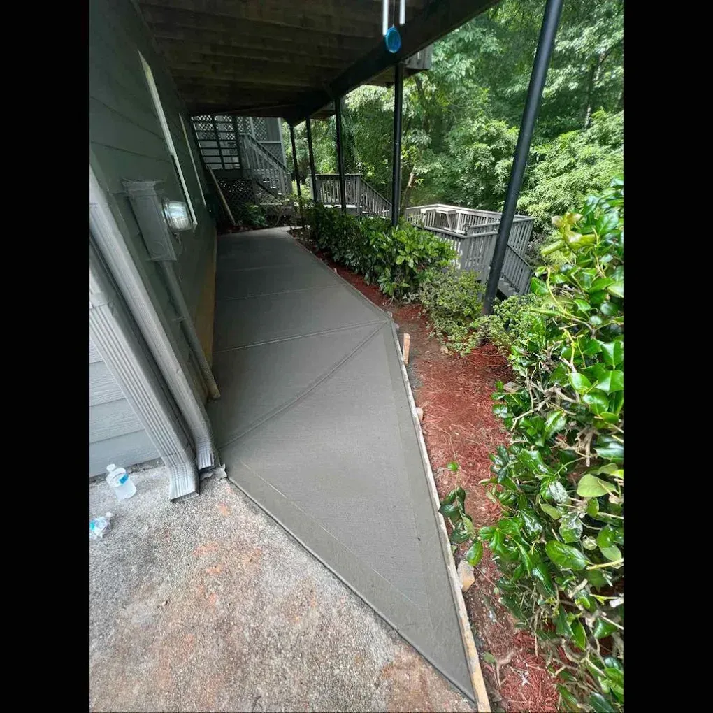Newly poured concrete walkway next to a building and landscaping, under a porch.