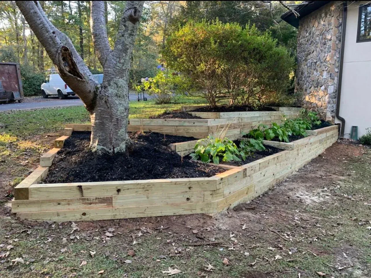 Wooden tiered garden beds surrounding a tree, with plants and dark mulch, beside a house.