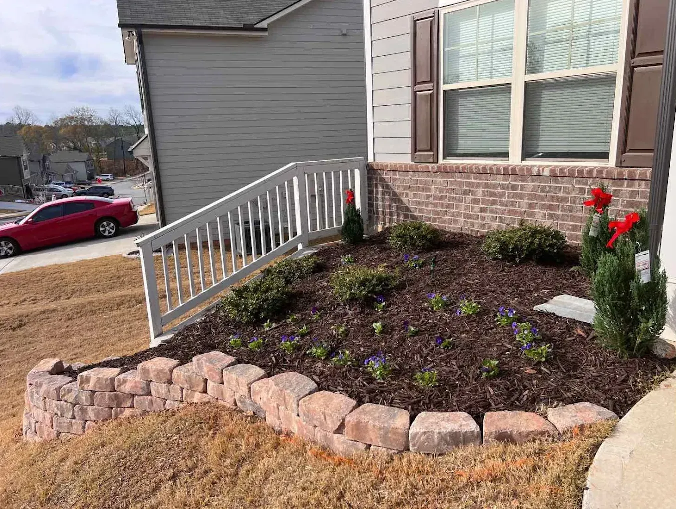 A small brick retaining wall and a flower bed beside a house with white railings and red car.