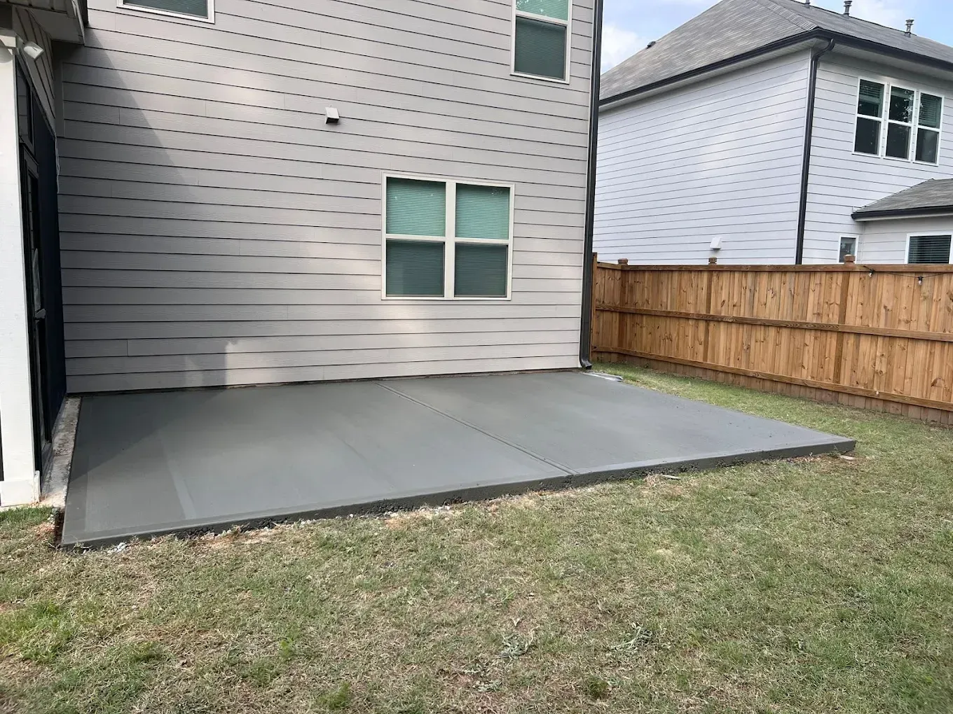 Concrete patio outside a two-story house with gray siding, next to a wooden fence and green grass.