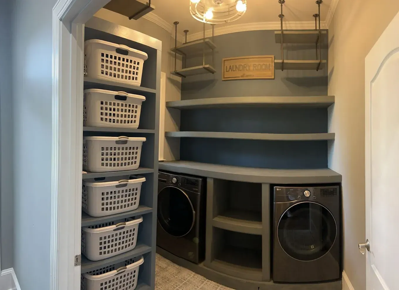 Laundry room with washing machine, dryer, shelves, baskets, and a sign. Gray and white color scheme.