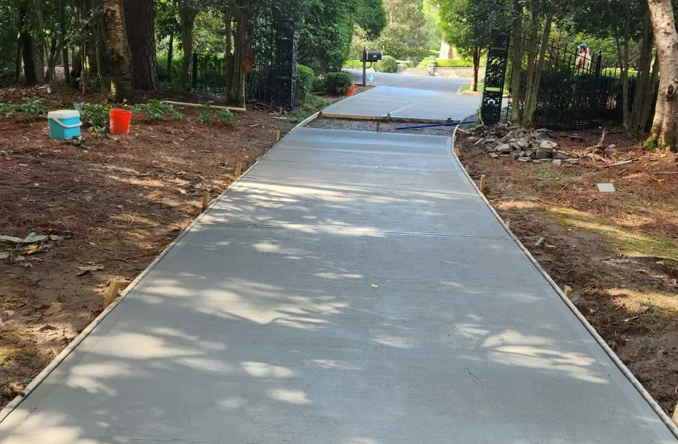 Newly poured concrete walkway winding through a wooded area; two buckets, red and blue, visible in the left.