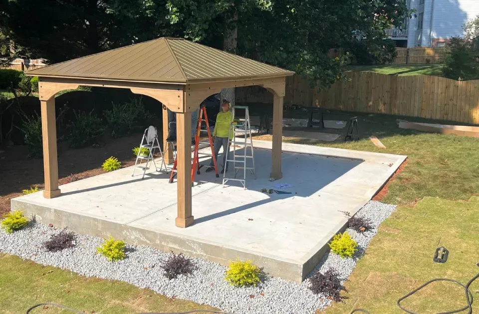 Workers constructing a brown gazebo on a concrete patio, surrounded by landscaping and a wooden fence.