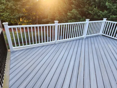 Gray wooden deck with white railing, bathed in sunlight, overlooking trees.