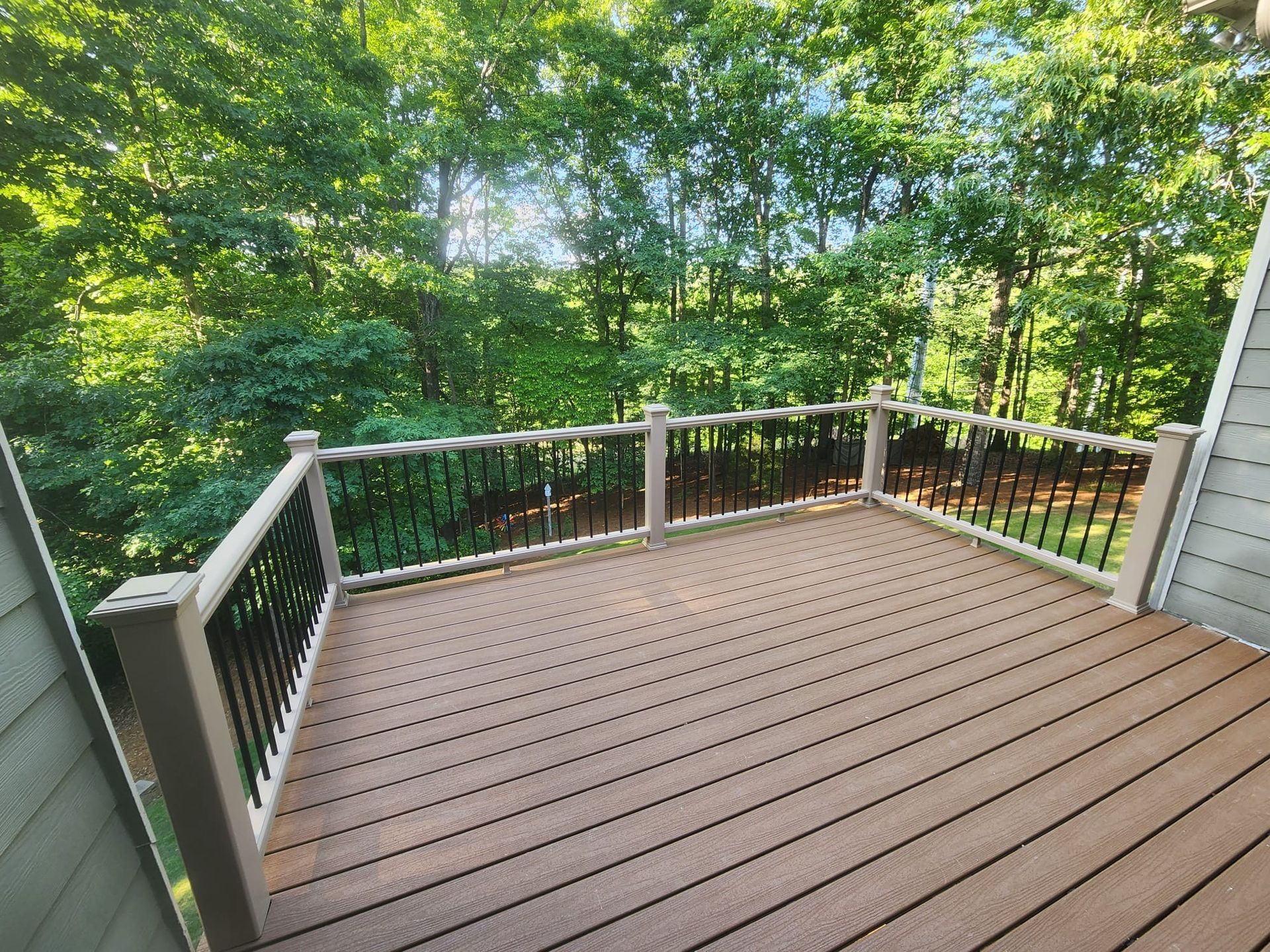 Deck overlooking a wooded area, brown decking, black railing, white posts, and green trees in the background.