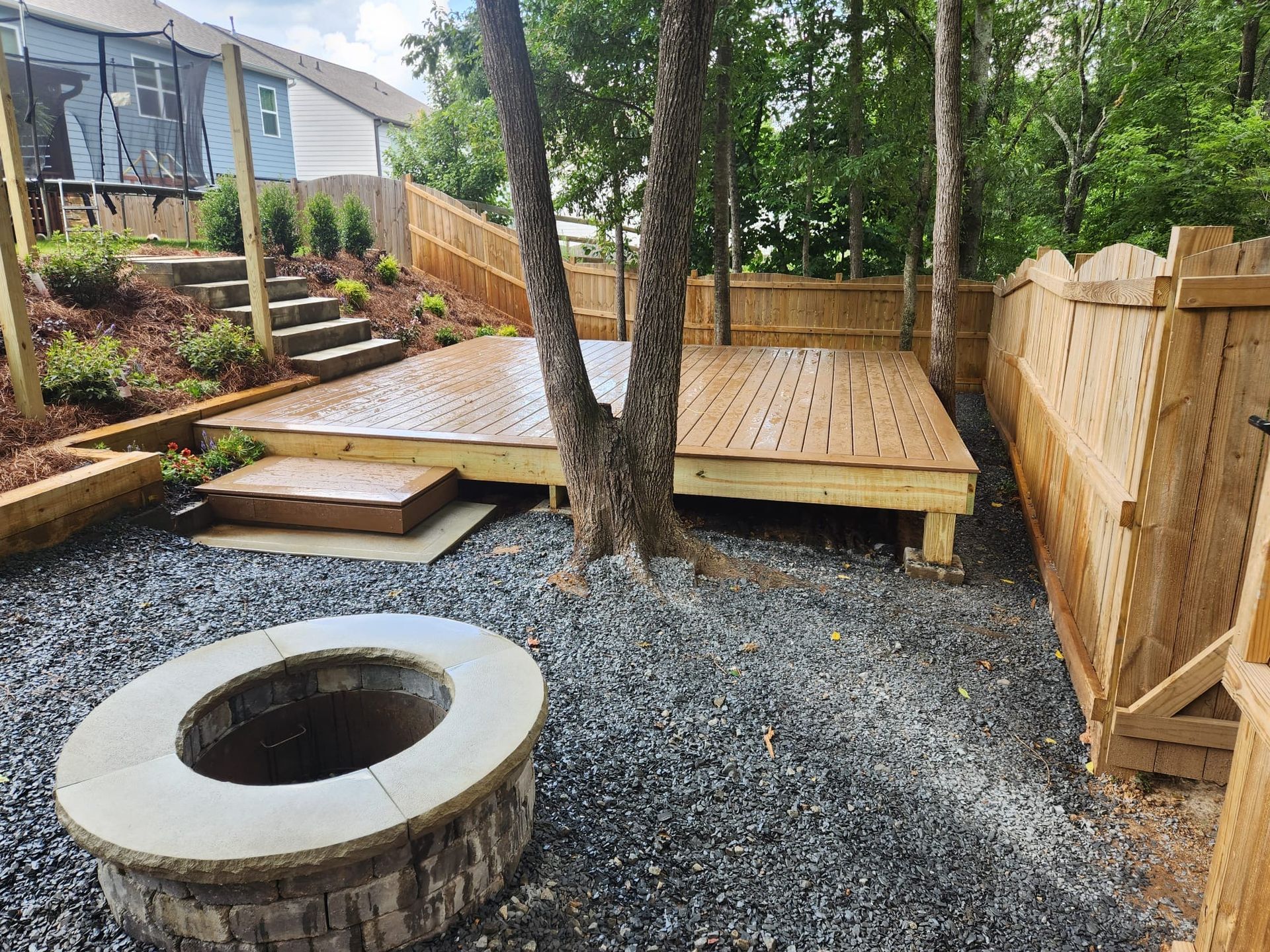 A backyard with a fire pit, wooden deck, and gravel ground; a fence and trees are present.