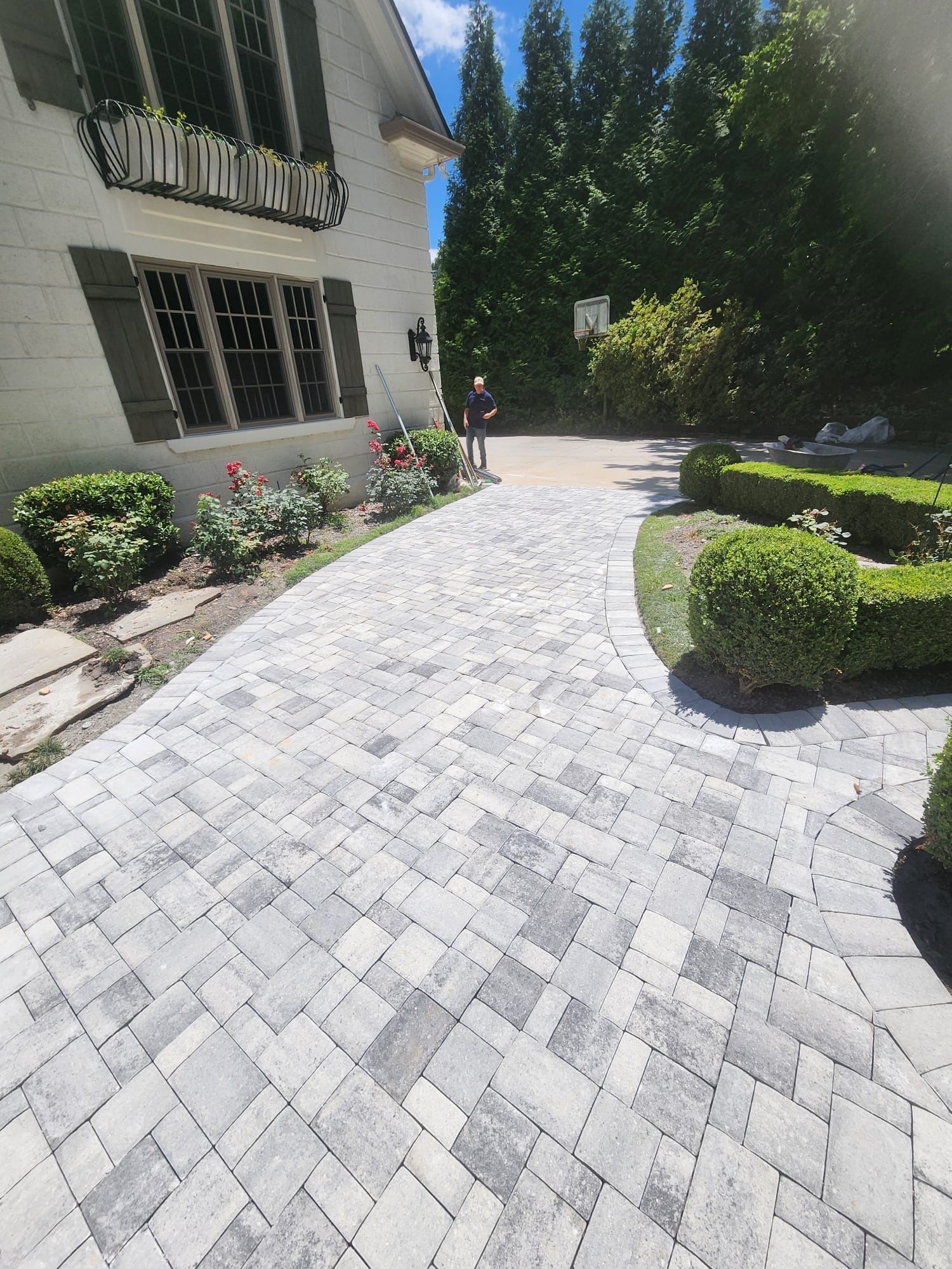 Brick pathway leading to a house, bordered by bushes and flowers. A person stands near the driveway.