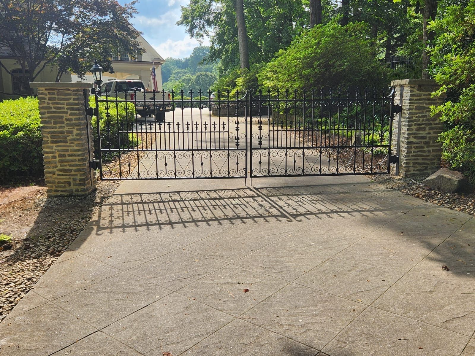 Black wrought iron gates between stone columns on a concrete driveway, leading to a house.