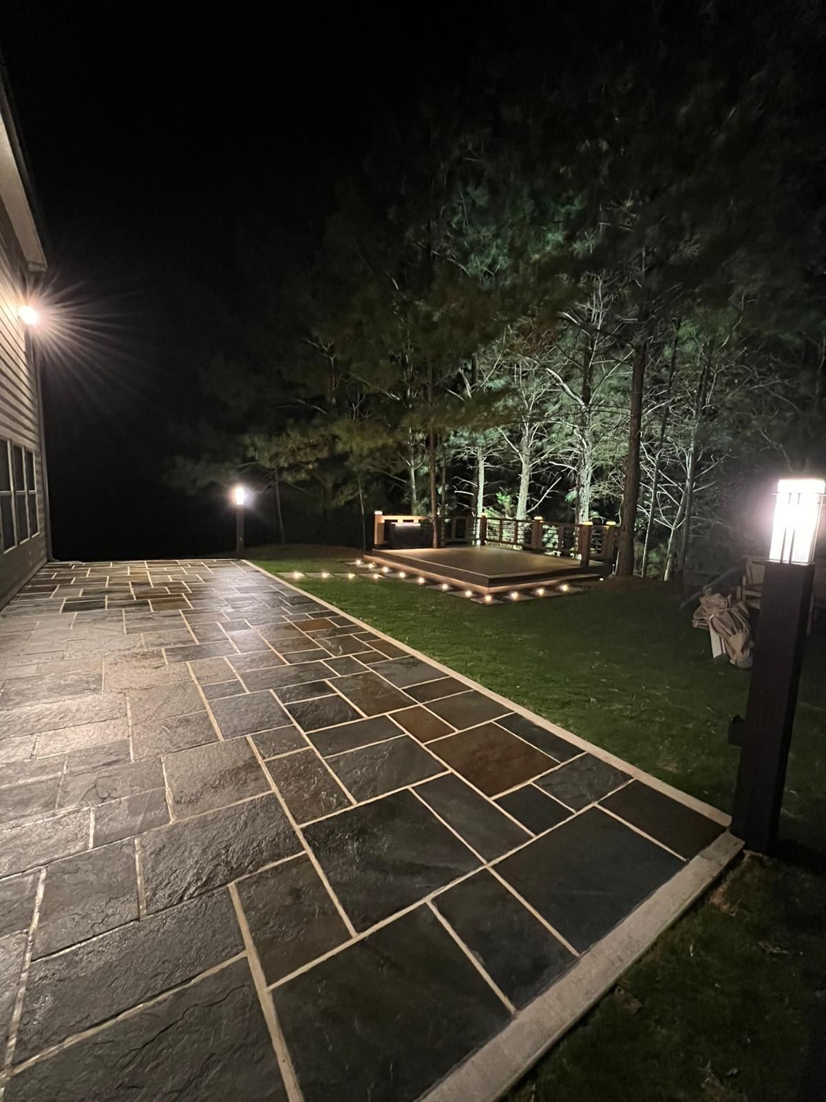 Night view of a patio with pathway, lawn, and lit stage under trees.