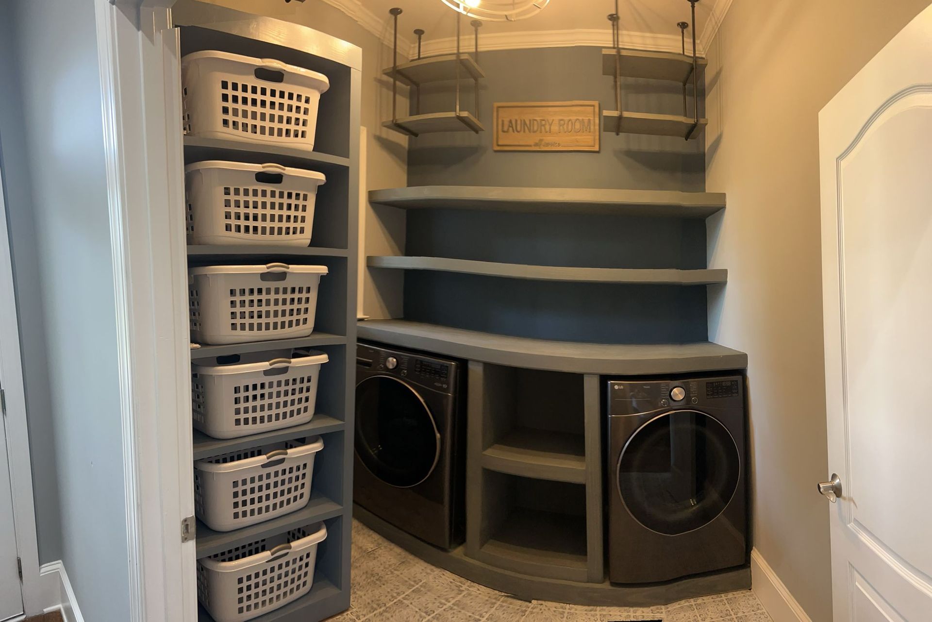 Laundry room with gray-painted shelves, washing machines, and six laundry baskets on left.