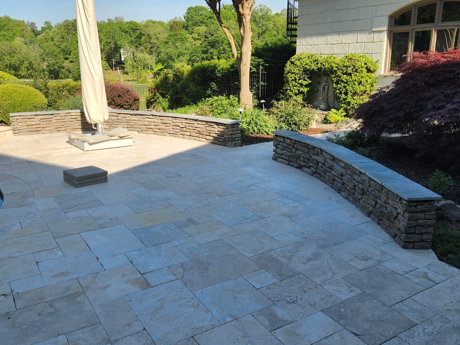 Stone patio with curved stone wall, plants, and an umbrella in a garden setting.