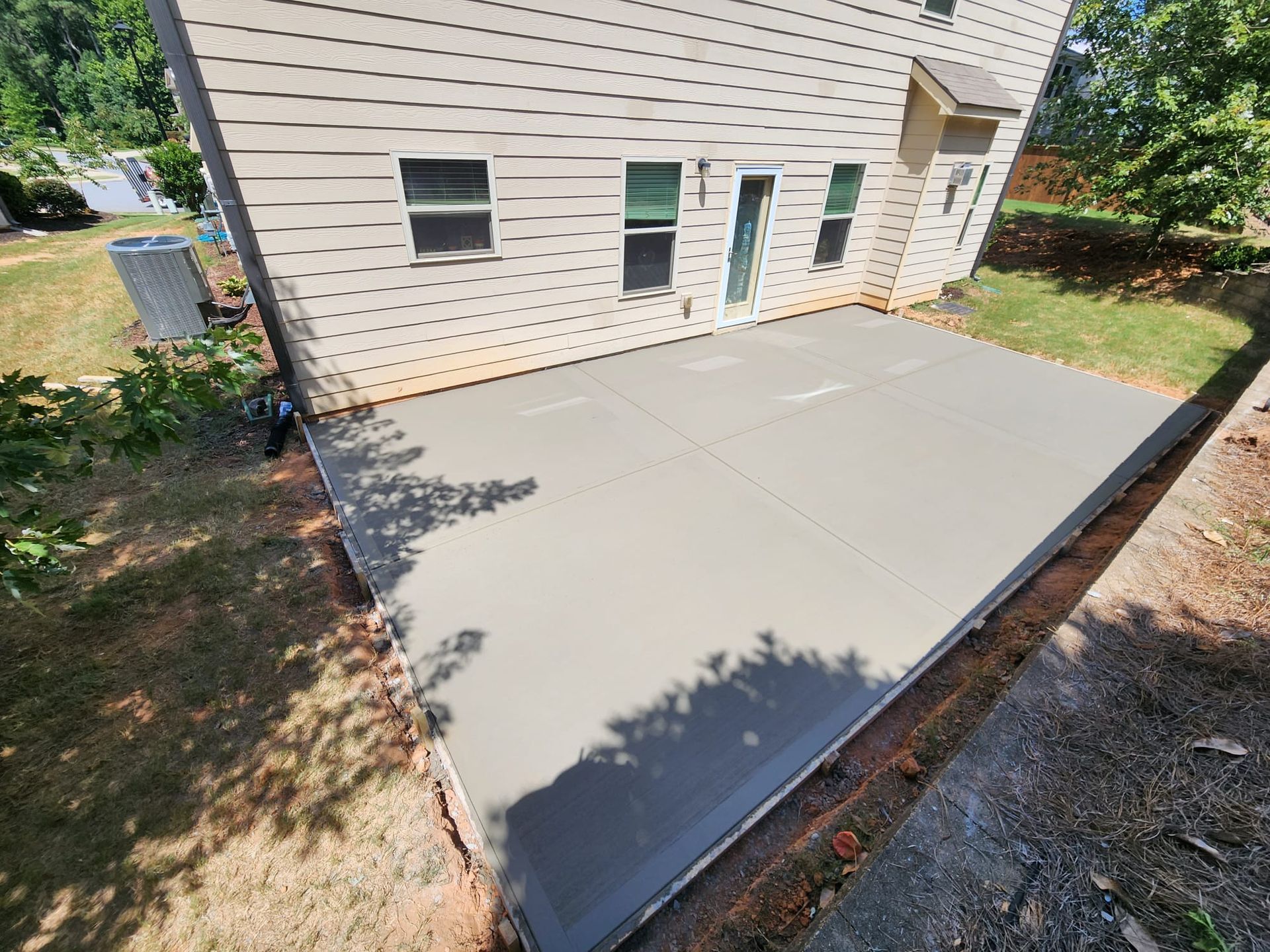 A freshly poured concrete patio adjacent to a light-colored house, surrounded by grass and red soil.