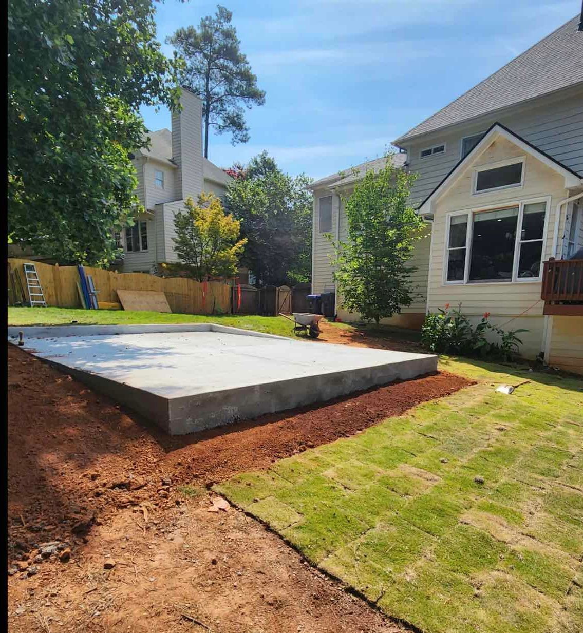 Concrete foundation in backyard with landscaping. Sunny day, green grass, dirt and surrounding homes.