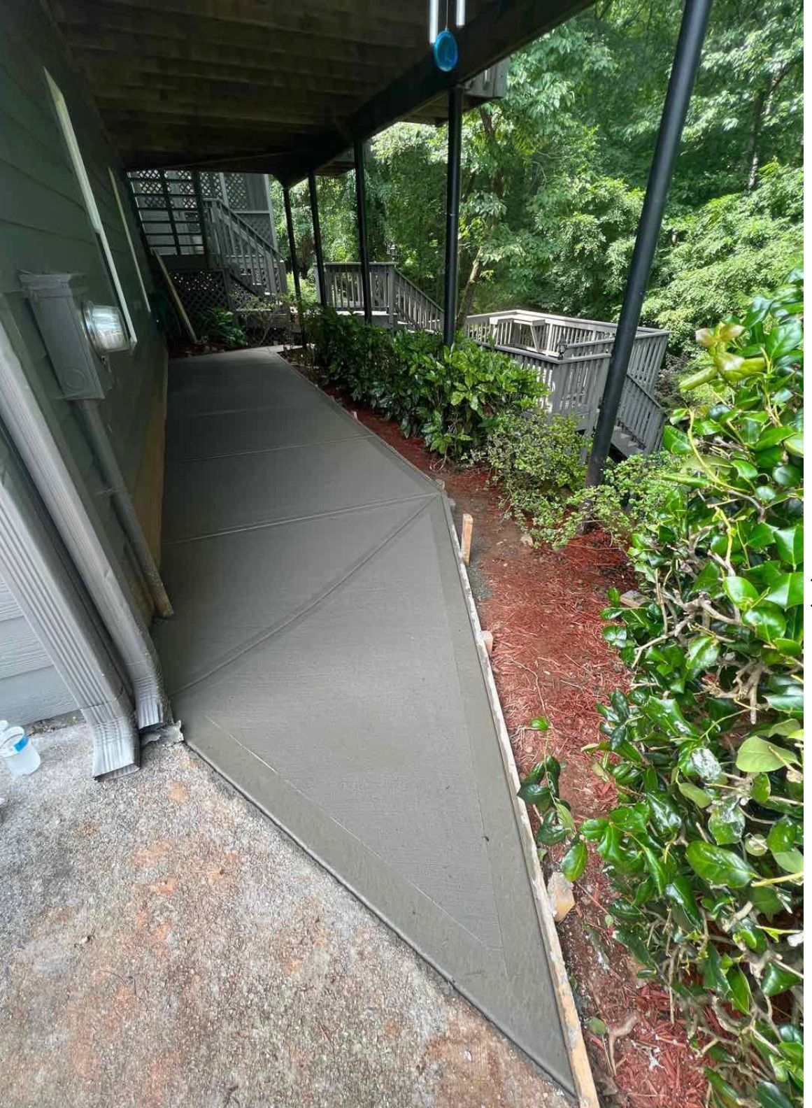 Concrete walkway with new pour, bordered by mulch and greenery, under a covered deck.