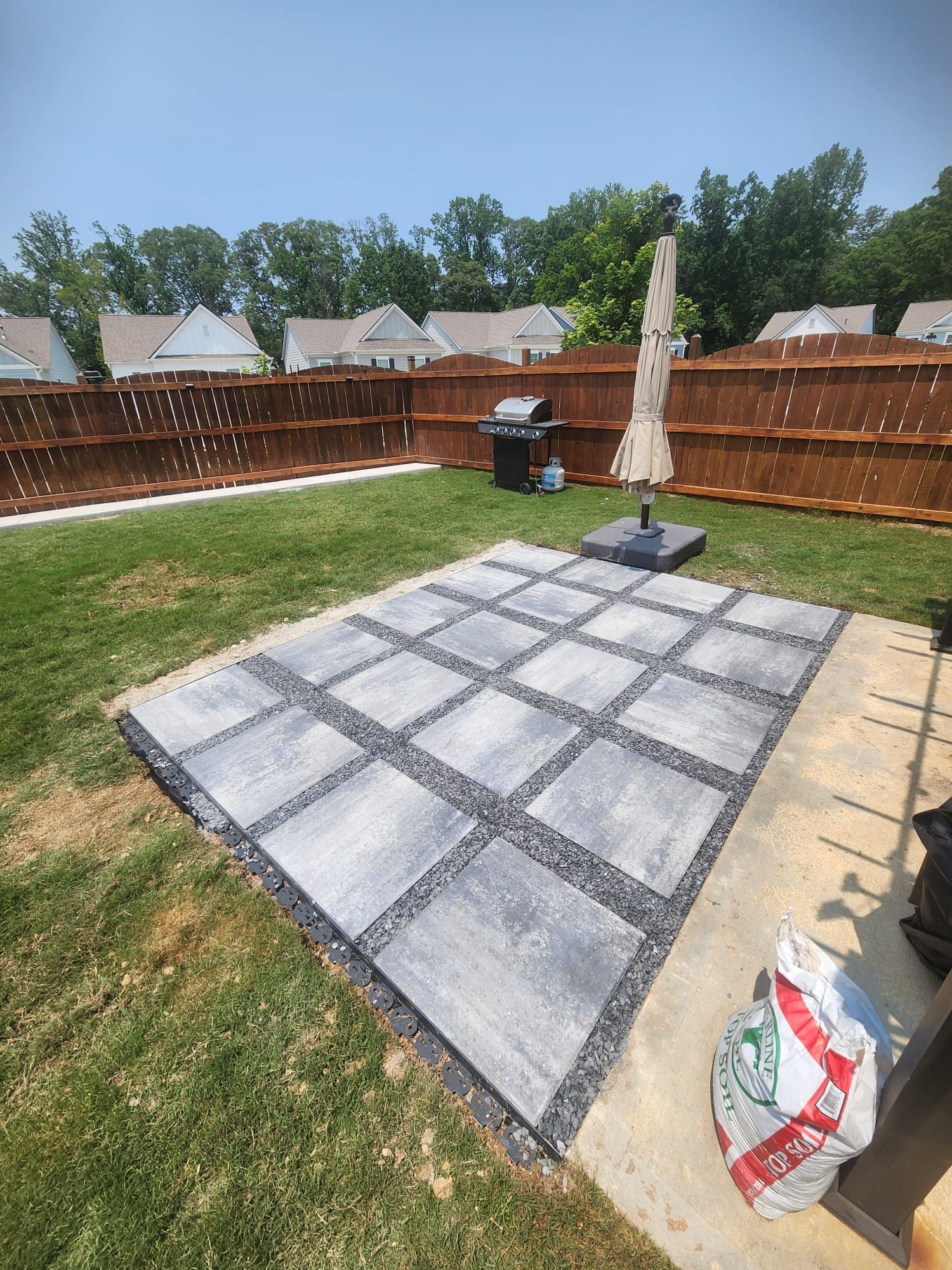 Backyard patio with gray pavers and dark gravel, grill, umbrella, and wooden fence.