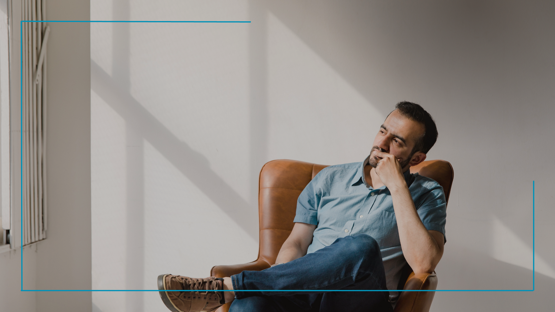 Man in blue shirt and jeans, sitting in brown chair, hand to face, looking up thoughtfully. Light from window.