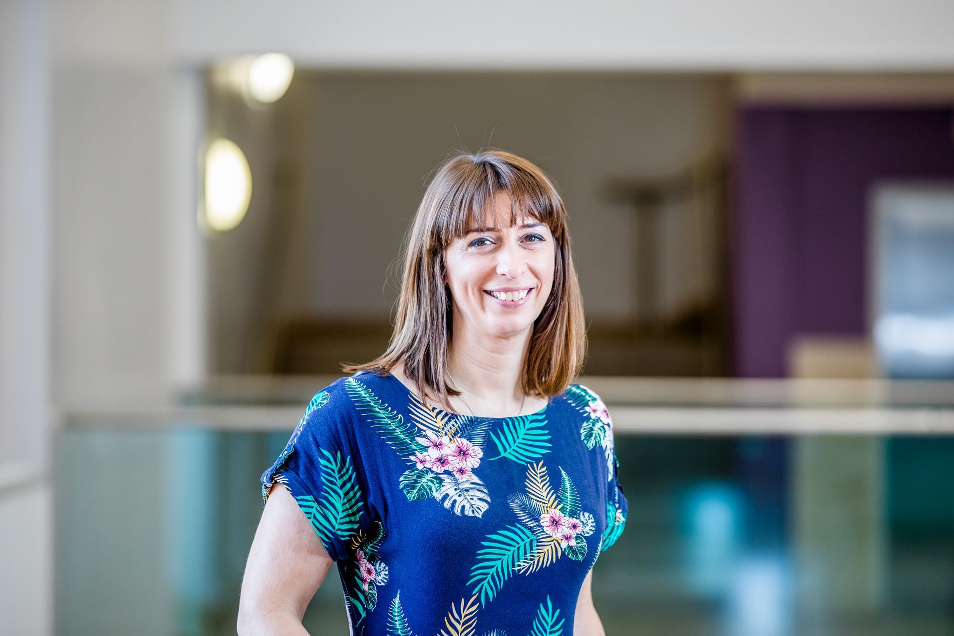 Woman smiling in blue floral top, standing in a brightly lit building.