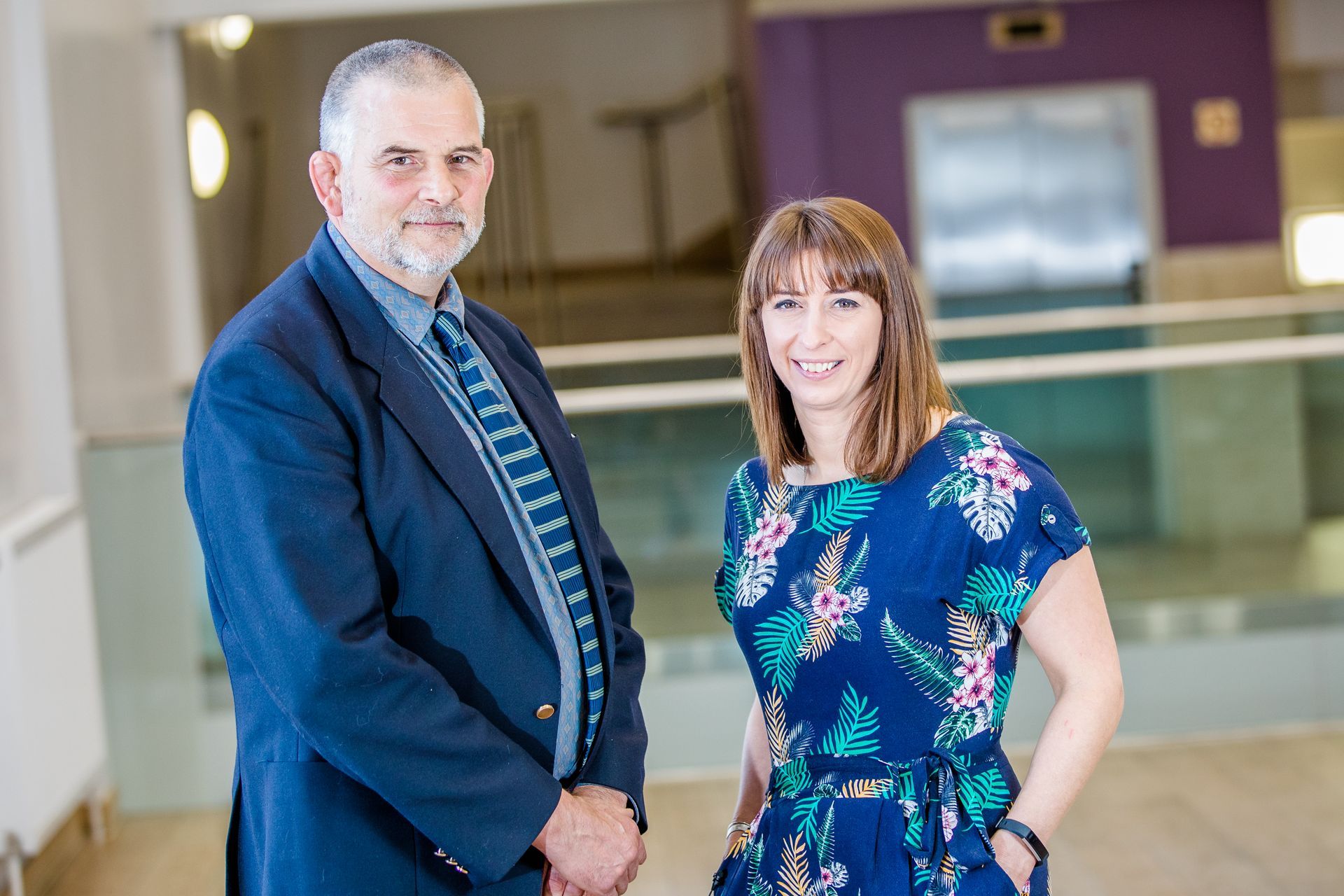 Man and woman standing indoors; man in a blue blazer, woman in a floral dress.