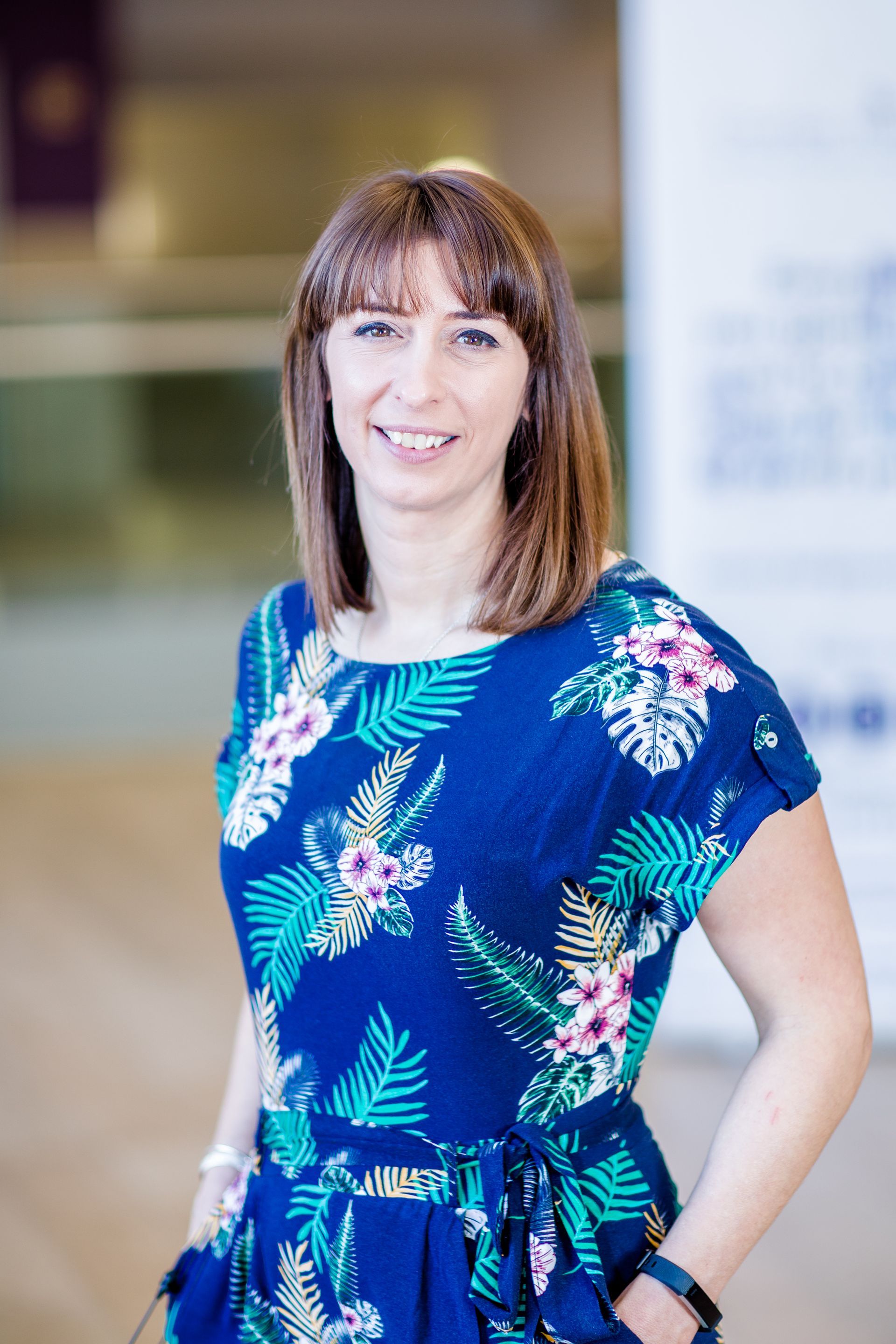 Woman in blue floral dress smiles, hands in pockets. Neutral indoor background.