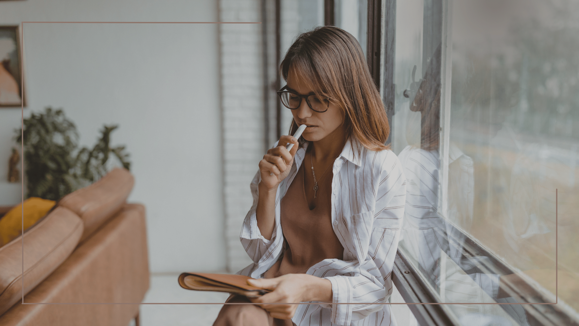 Woman in glasses and white shirt sits by window, holding pen to her lips and looking at notepad.