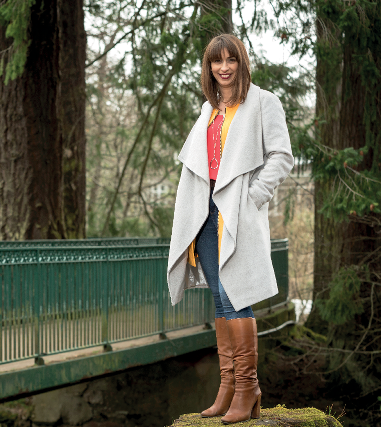 Woman standing on mossy rock, wearing gray coat, orange top, jeans, brown boots, in wooded park by a bridge.