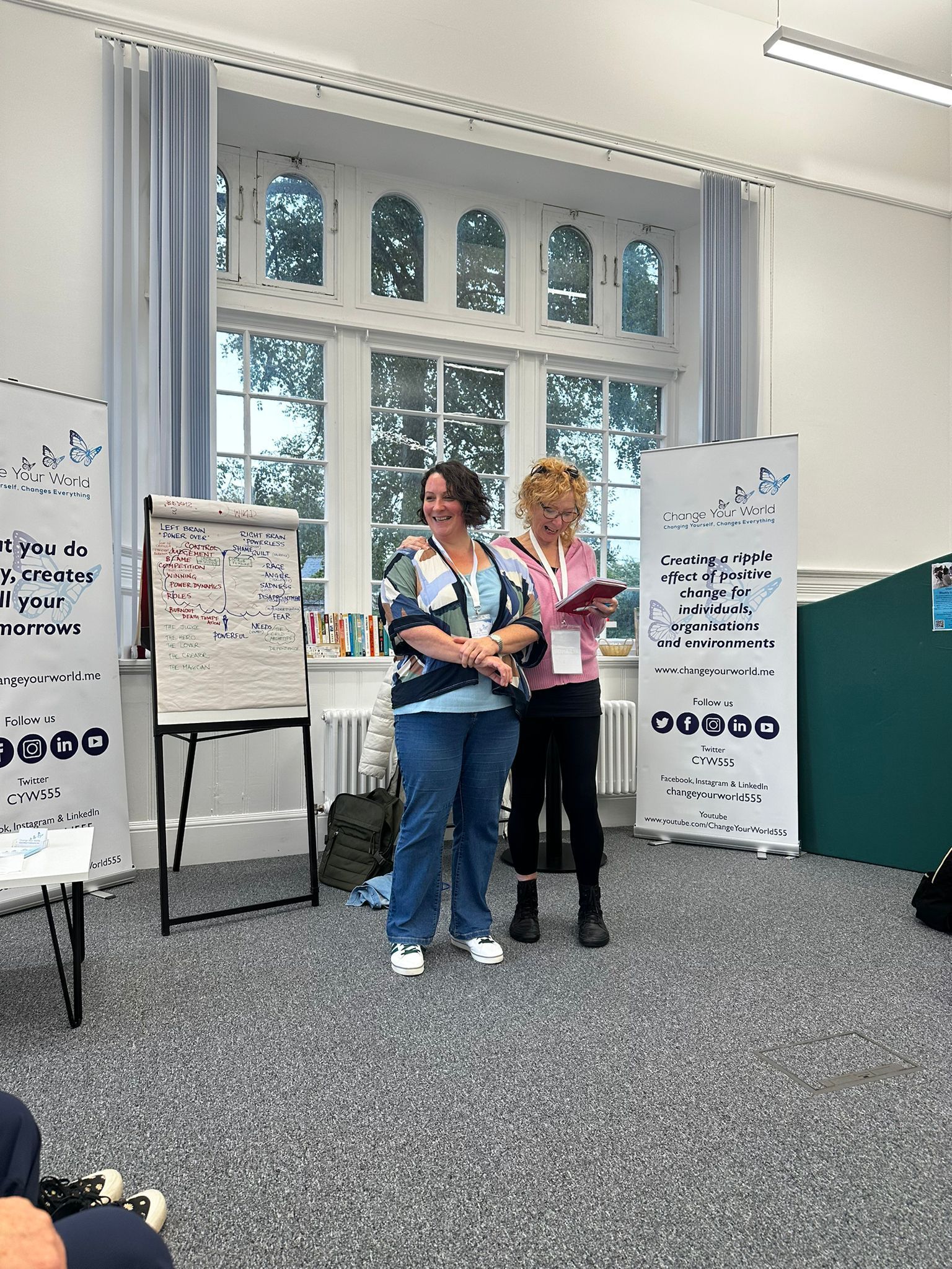 Two women stand indoors in front of a window; one holds an award. A whiteboard and banners are visible.