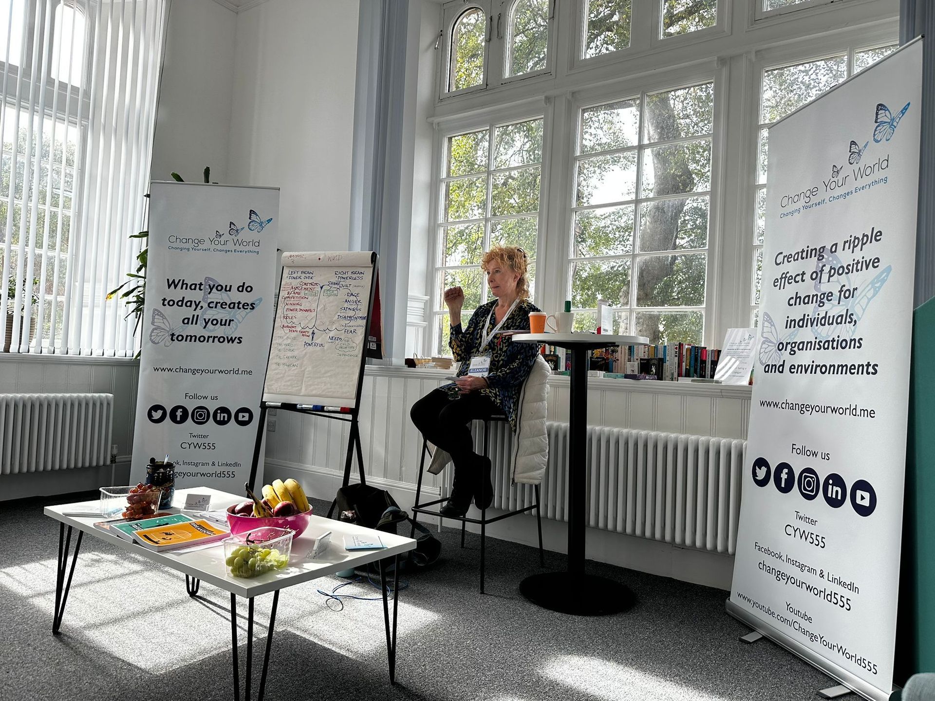A woman sits in front of a window, two banners behind her. She's at a table with fruit and snacks.