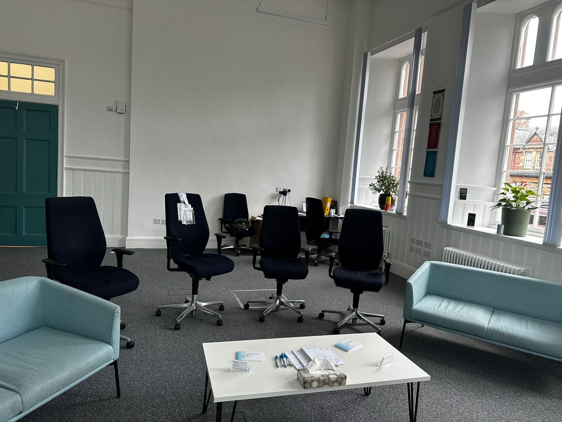 An office meeting room with blue chairs, a couch, and a white table. Windows and a green door are visible.