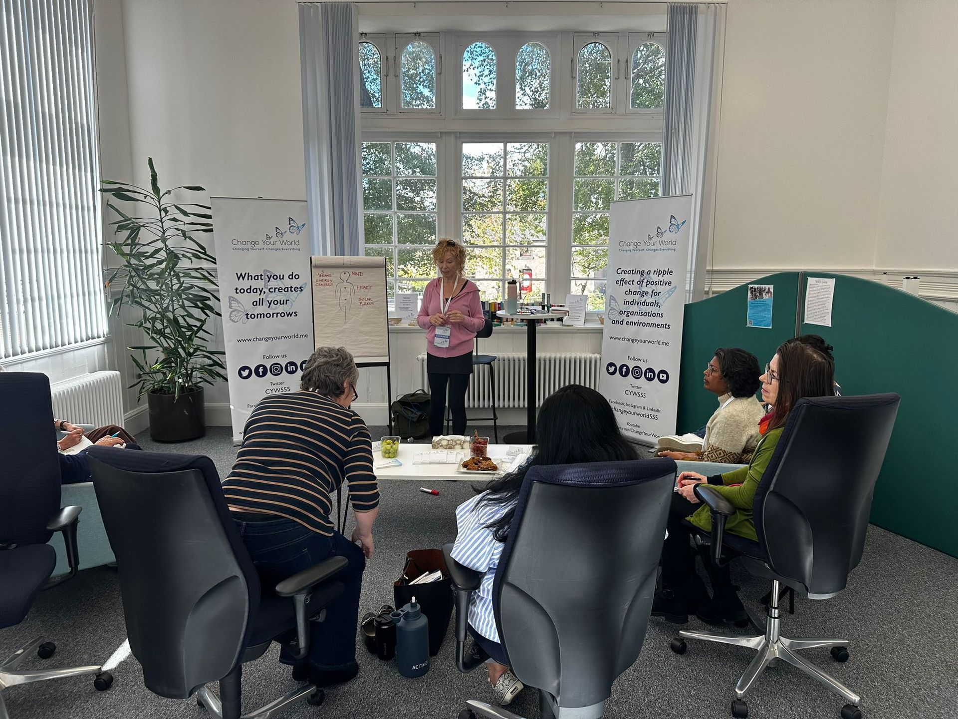 A person giving a presentation in a room with a window, banners, and people seated around a table.