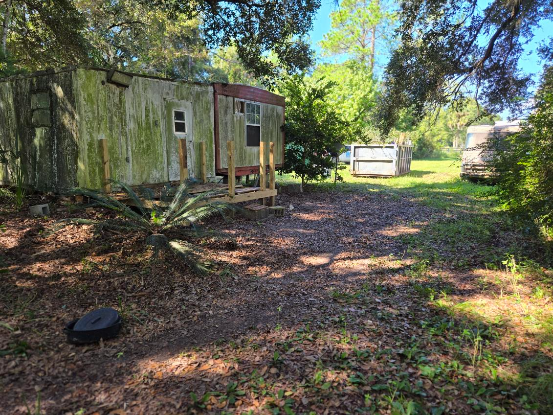 Dilapidated trailer in a wooded lot; overgrown vegetation and two other trailers visible.