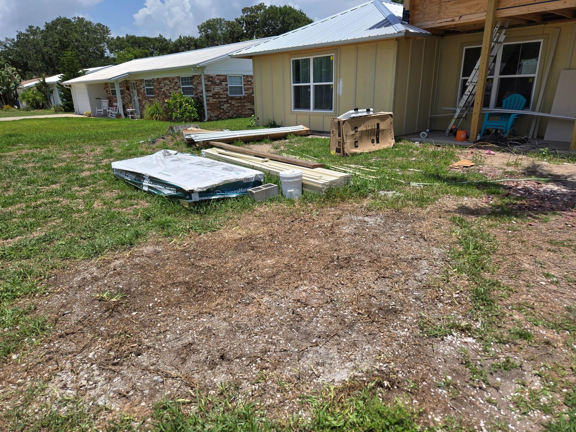 Construction materials scattered on a grassy yard next to a yellow house with a metal roof.