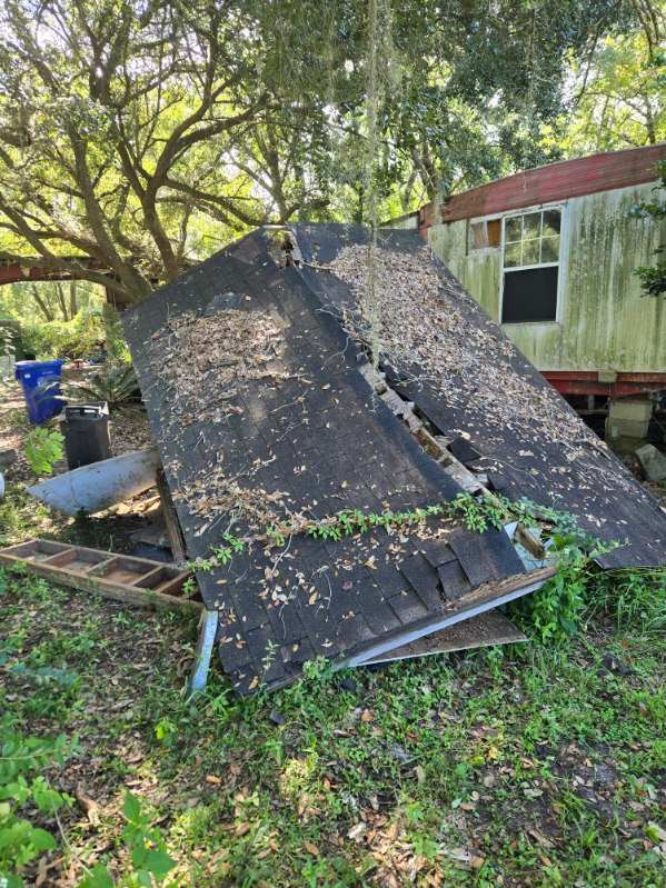 Fallen roof shingles lie on ground next to a rundown building in a grassy area with trees.