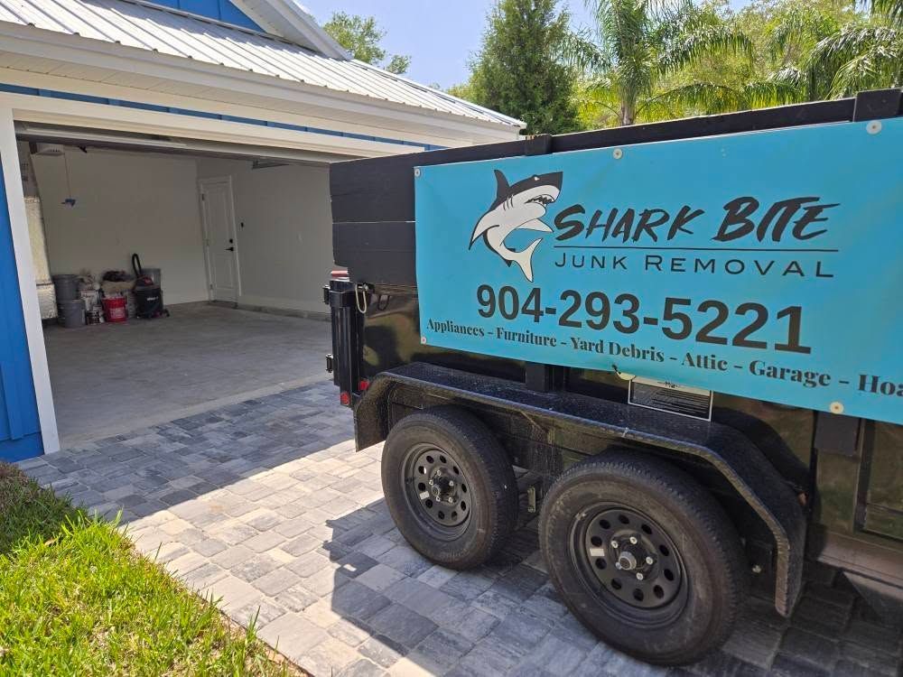 A shark bite junk removal trailer is parked in front of a garage.
