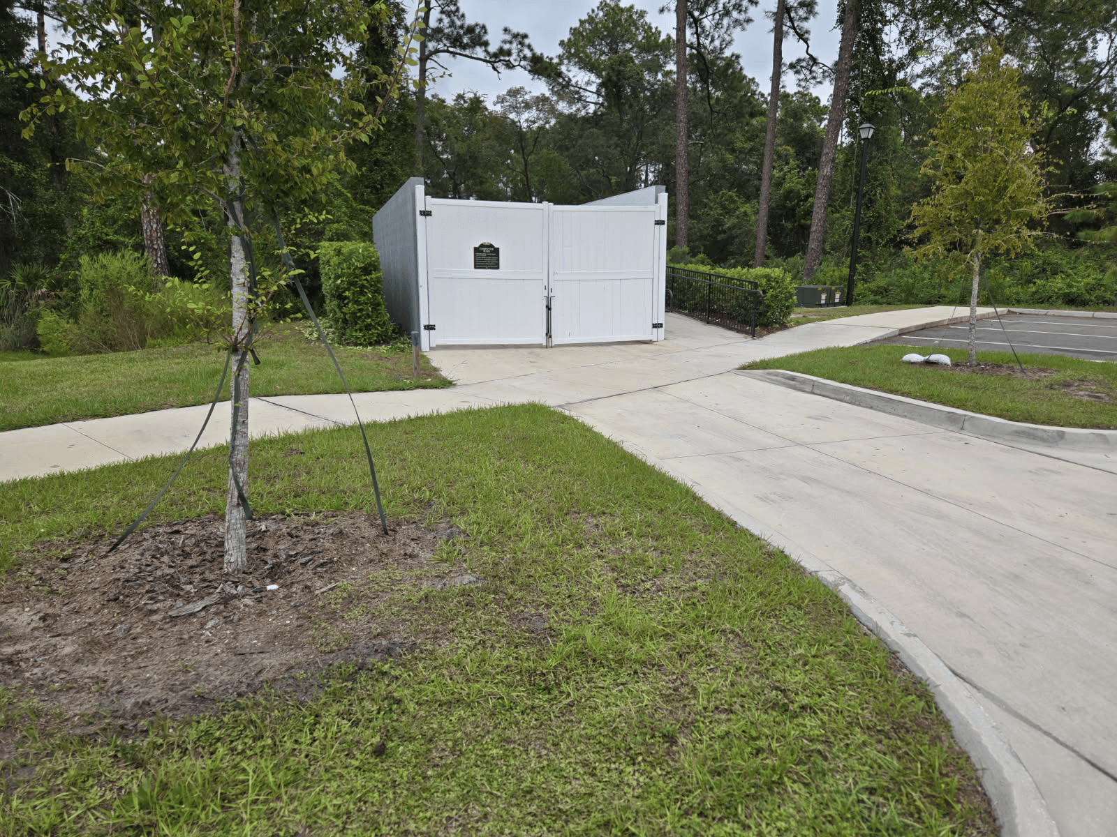 White gated storage building at the end of a concrete driveway, surrounded by green grass and trees.