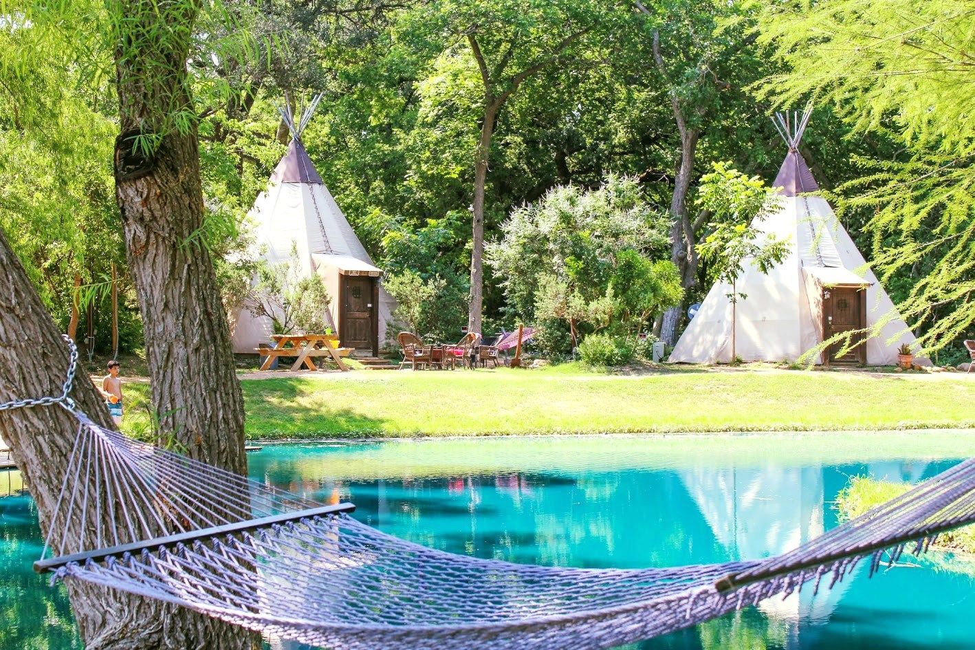 A hammock is hanging from a tree next to a lake with teepees in the background.