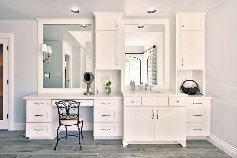 A bathroom with white cabinets , a vanity , two mirrors and a chair.