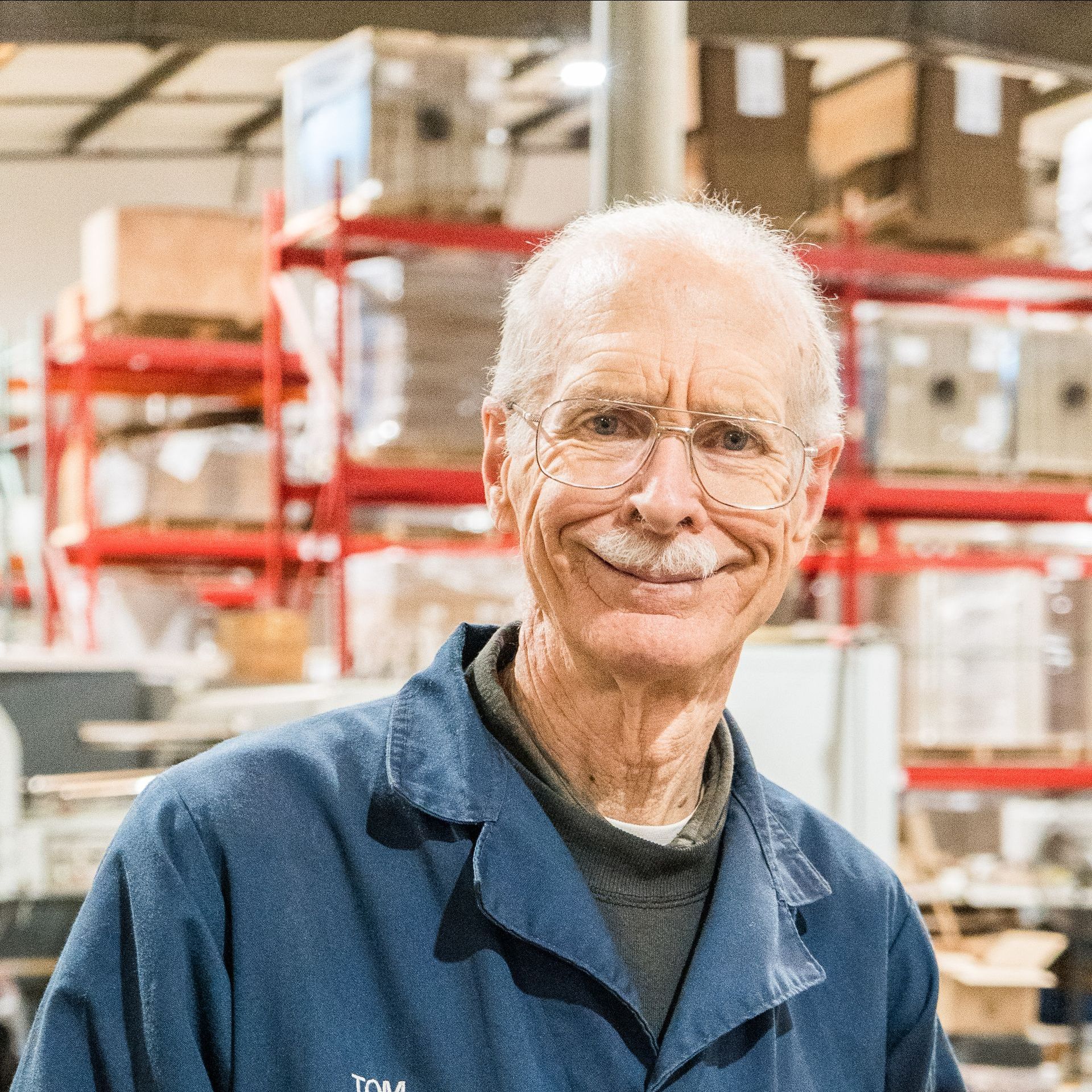 An older man wearing glasses and a blue jacket is smiling in a warehouse.