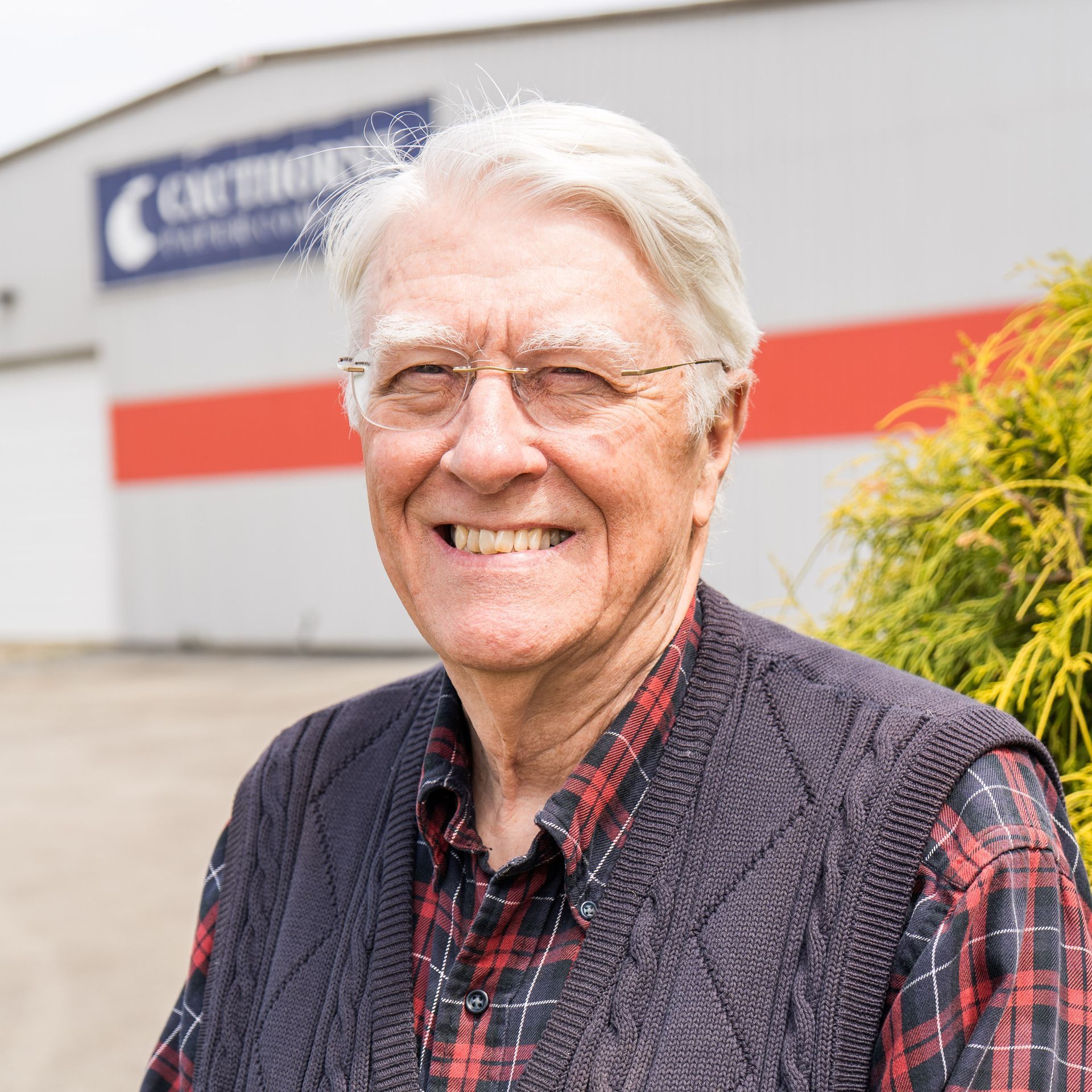 An older man wearing glasses and a plaid shirt is smiling in front of a building.