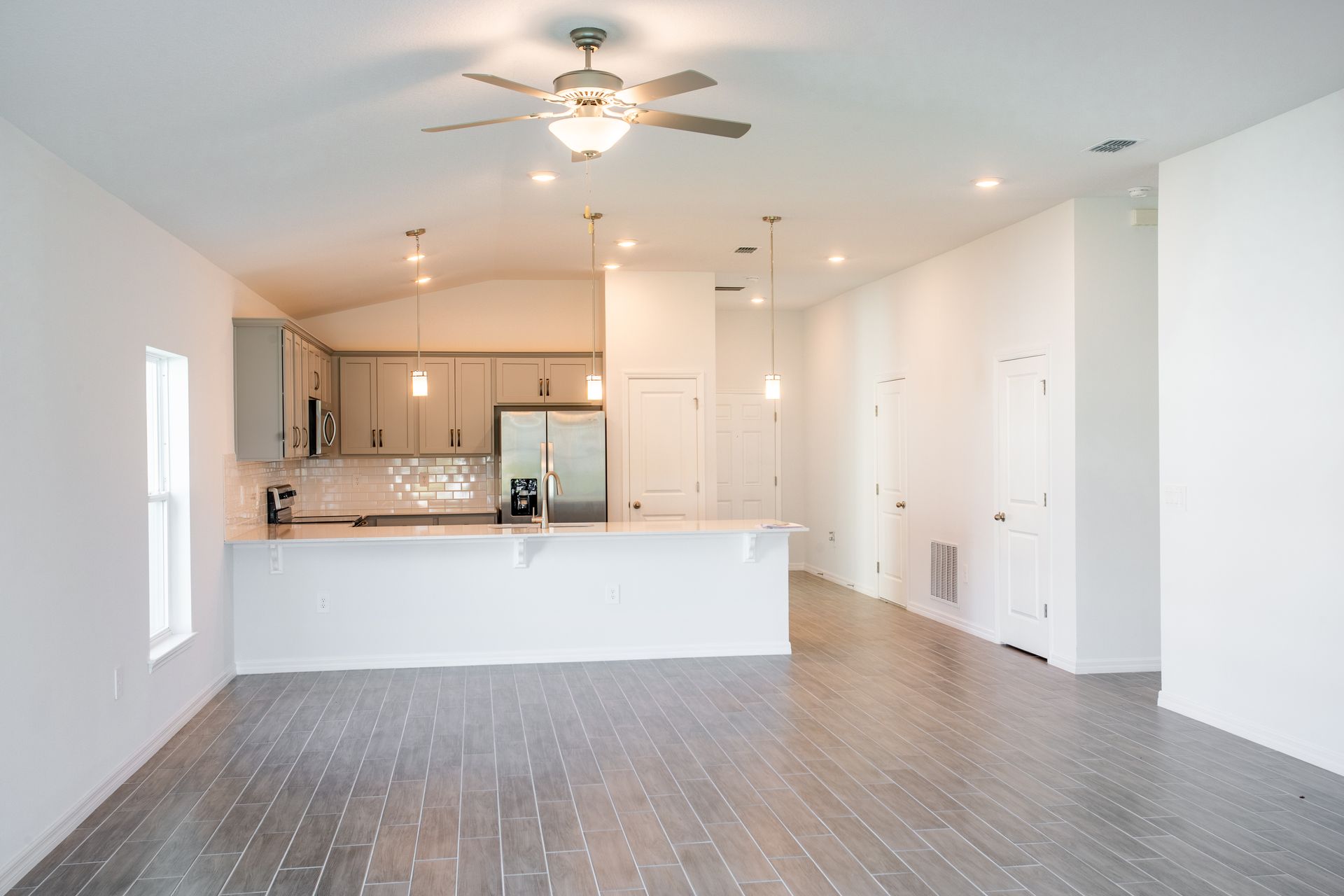 An empty living room with a ceiling fan and a kitchen in the background.