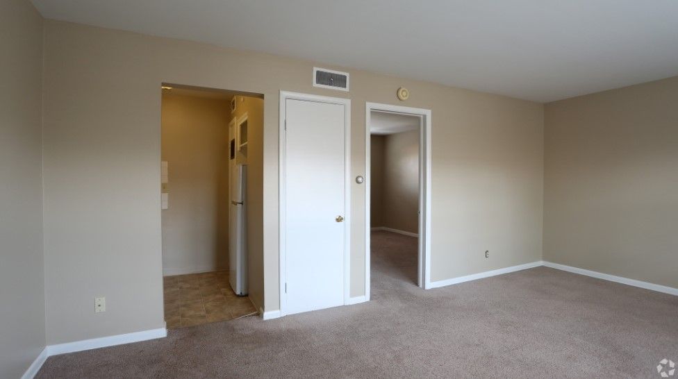 An empty living room with a carpeted floor and a refrigerator.
