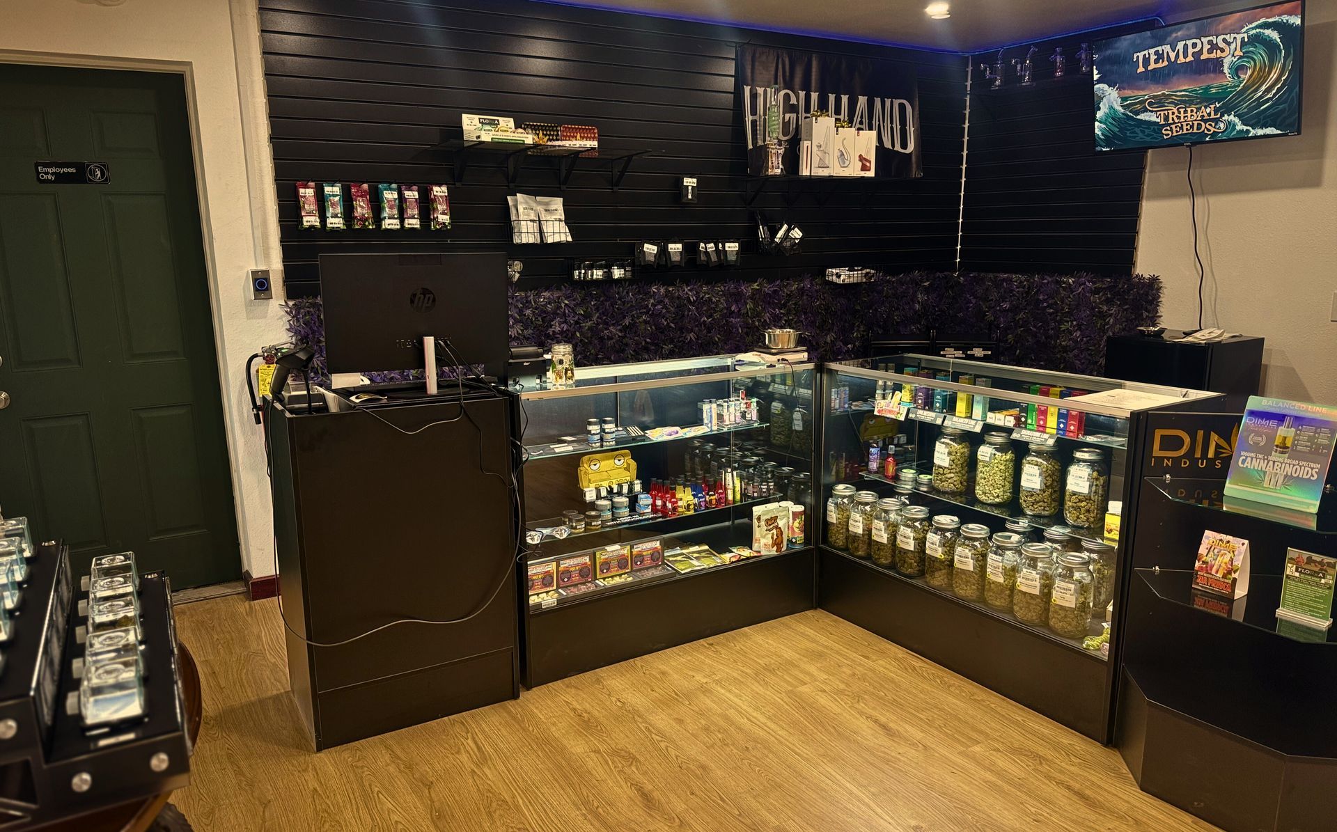 Interior of a retail shop displaying cannabis products, with glass display cases, jars, and a TV screen.