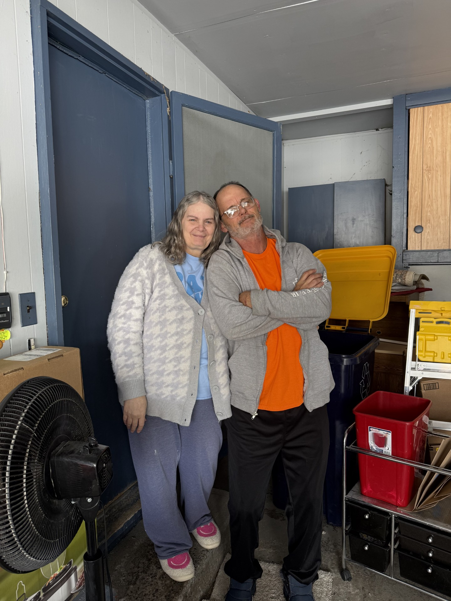 Couple standing inside, woman in sweater, man with arms crossed, next to a blue door.