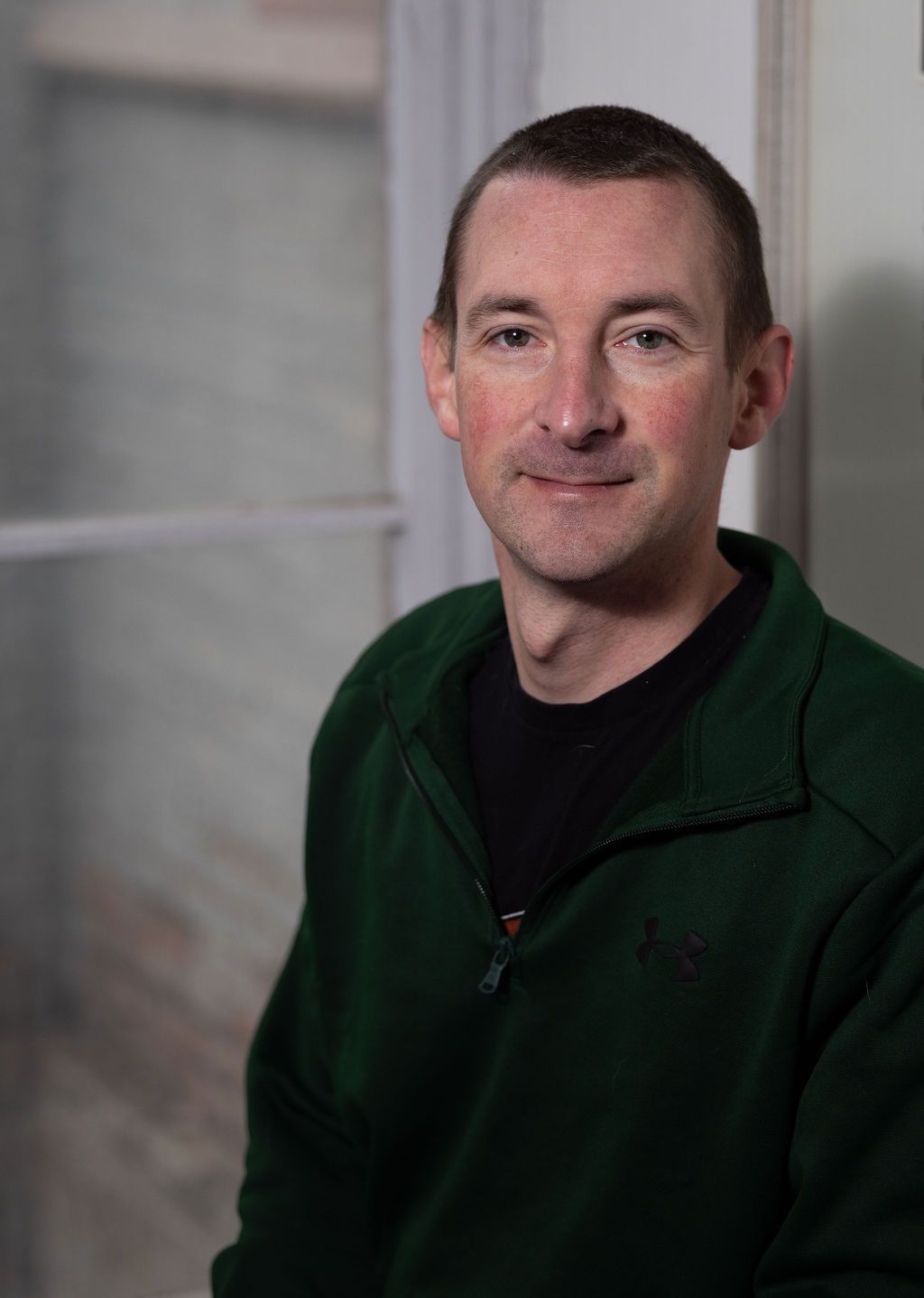 Man in green sweater smiles in front of a window.