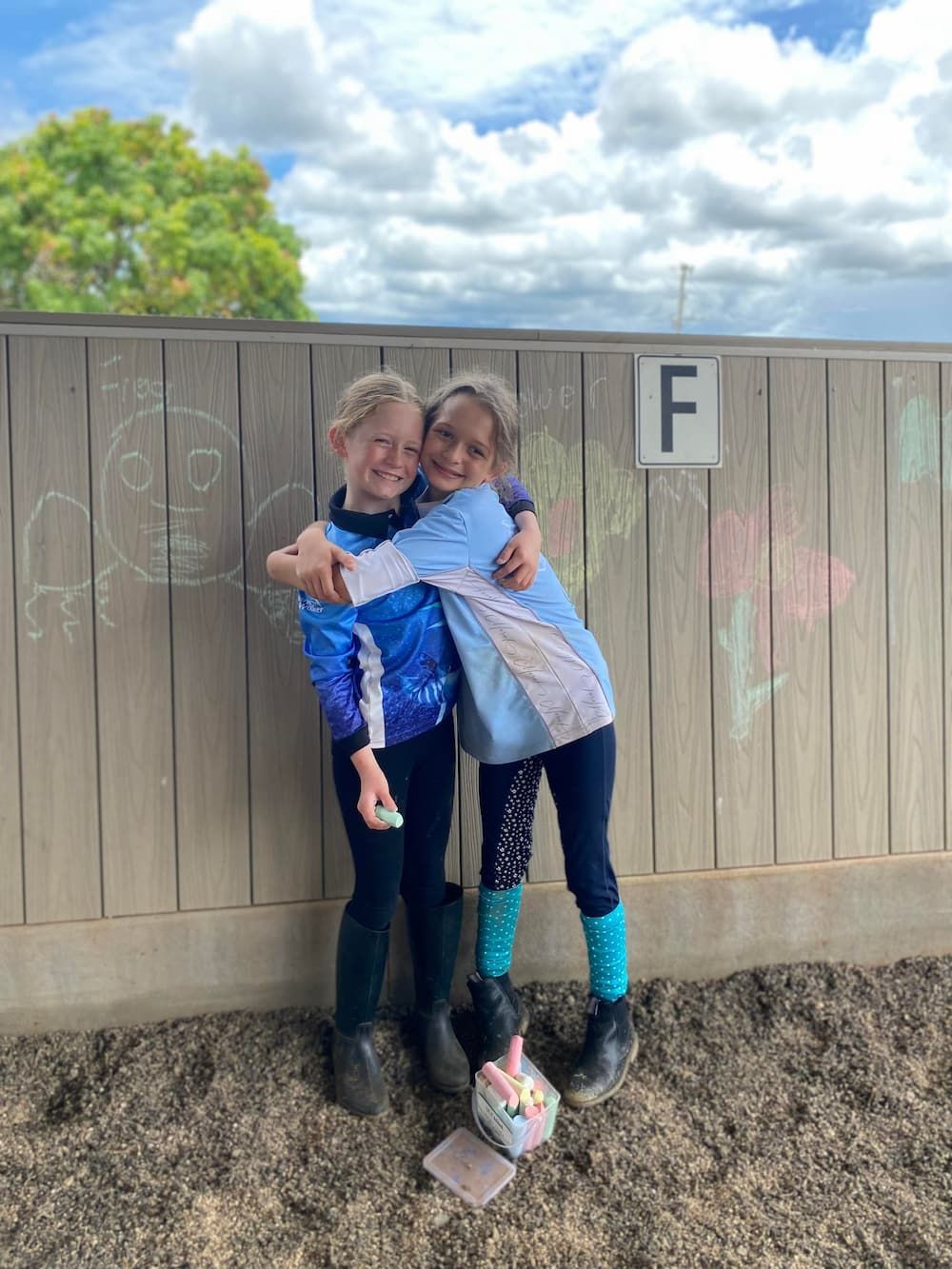 Two Young Girls Are Hugging Each Other in Front of a Wooden Fence — Meraki Equestrian Centre in Mareeba, QLD