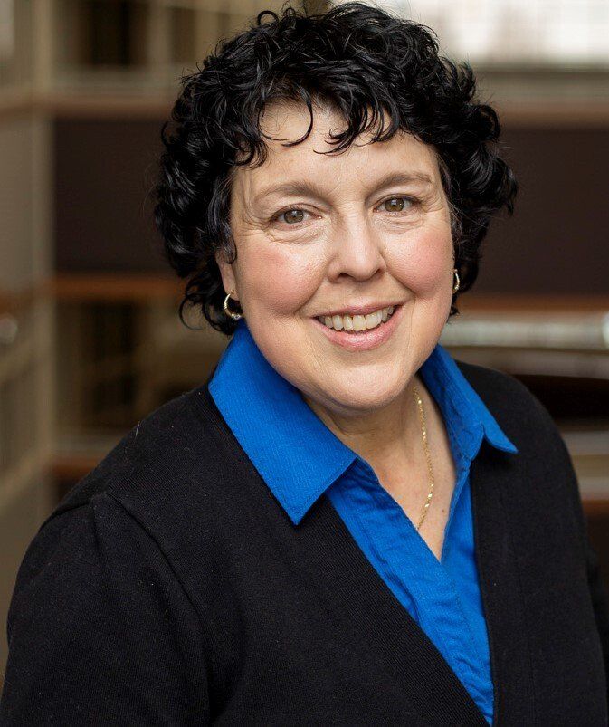 A woman wearing a blue shirt and a black sweater smiles for the camera